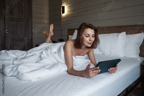 Girl lying in bed using digital tablet In bedroom. Woman lying in bed in hotel room using mobile device for work. Young girl on vacation hotel room on white bed. Person is working while in bed