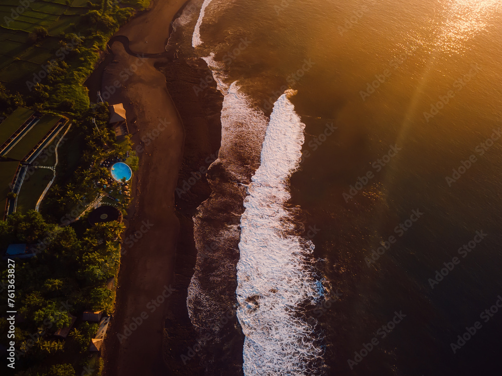 Aerial view of black sand beach with sunrise or sunset tones and ocean with waves in Bali