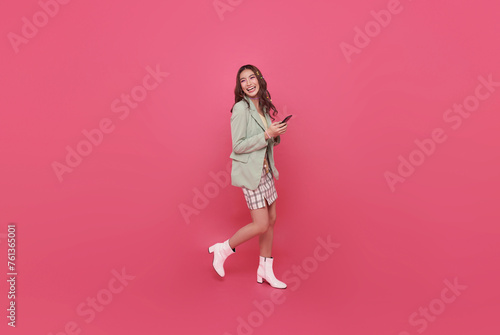 Young smiling happy asian woman 20s holding mobile cell phone and walking isolated on pink background. portrait People lifestyle concep