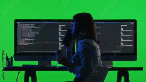 Back view of female programmer drinking coffee and answering phone call while coding on computer with dual monitor setup at desk in studio with green chroma key backgroundBack view of female programme photo