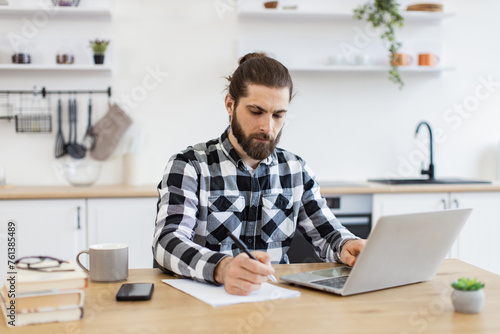 Handsome Caucasian man wearing checkered shirt writing on document while staying at home during midday. Efficient business owner improving contract while checking data on portable computer.
