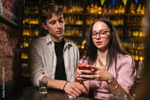 Guests spend relaxing time at bar counter in nightclub. Guy and lady talk with other comrades in remote format at party in club