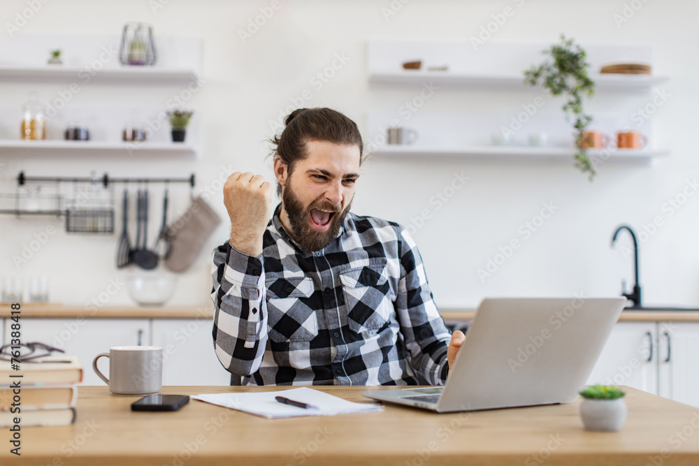 Emotional young businessman celebrating completion of great deal. Excited Caucasian adult wearing checkered shirt and jeans raising hands up looking at laptop happy of good results of work. Emotional young businessman celebrating completion of great deal. Excited Caucasian adult wearing checkered shirt and jeans raising hands up looking at laptop happy of good results of work.