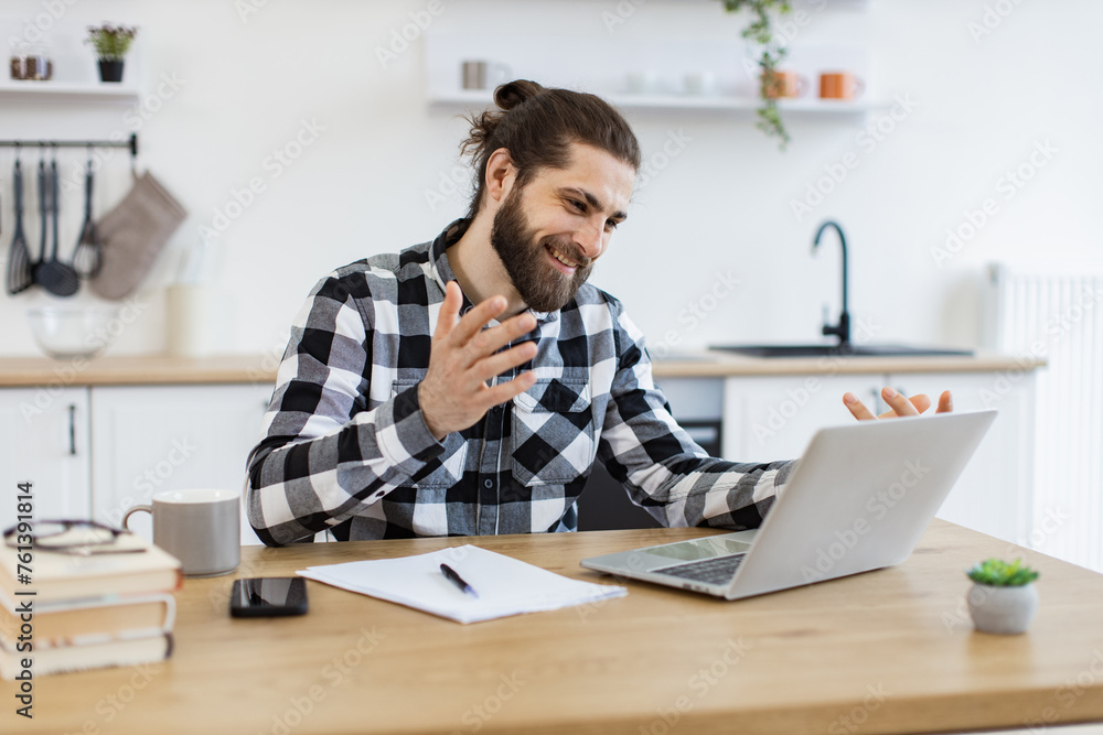 Naklejka premium Proficient Caucasian consultant receiving online conference call using technologies. Cheerful bearded adult looking at webcam of portable computer while greeting colleague in home office.