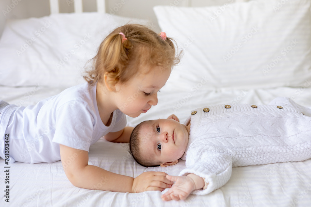 sister gently hugs and kisses a newborn baby in a crib on a white insulated cotton bed, two children in the family brother and sister sister gently hugs and kisses a newborn baby in a crib on a white insulated cotton bed, two children in the family brother and sister
