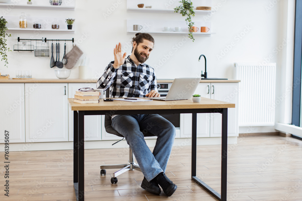 Cheerful bearded adult waving hello at webcam of portable computer while greeting colleague in home office. Proficient Caucasian consultant receiving online conference call using technologies.