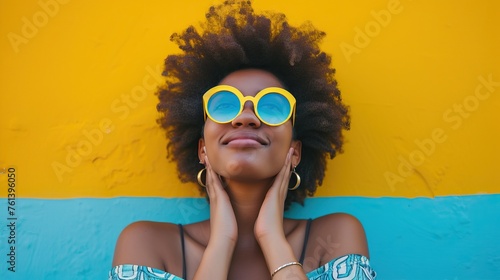 Young woman with afro hair wearing yellow sunglasses against a vibrant yellow wall, exuding happiness and style.