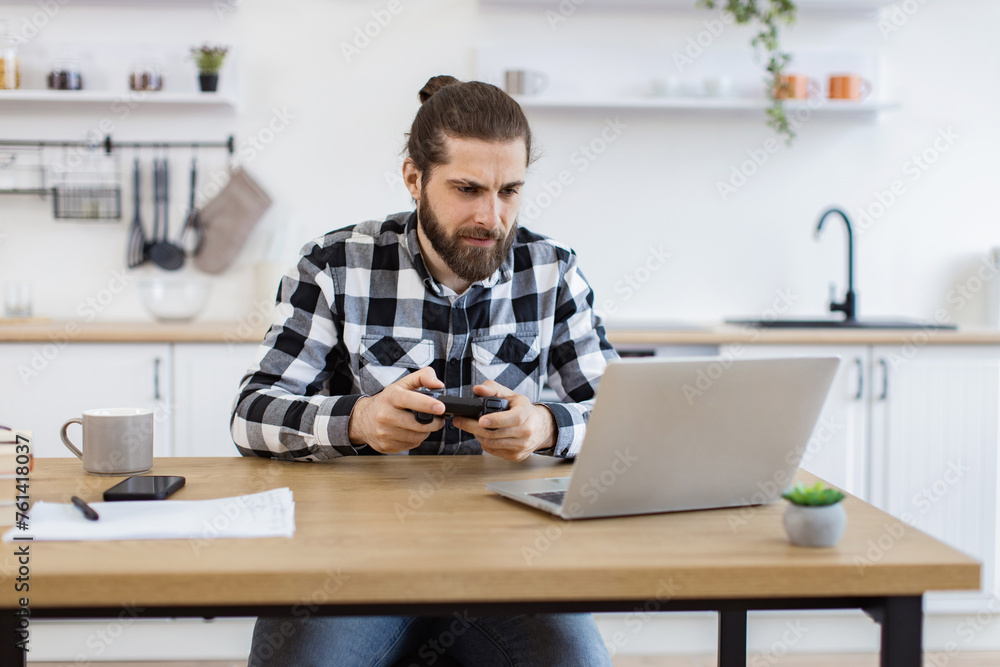 Portrait of young Caucasian man using wireless joystick and laptop sitting at wooden table at kitchen. Male freelancer taking break and plays video games while working during evening time. Portrait of young Caucasian man using wireless joystick and laptop sitting at wooden table at kitchen. Male freelancer taking break and plays video games while working during evening time.