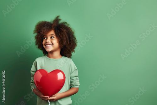 A kid model with a charming smile stands against a green solid wall, expressing love by holding a heart-shaped balloon and looking affectionately at it.