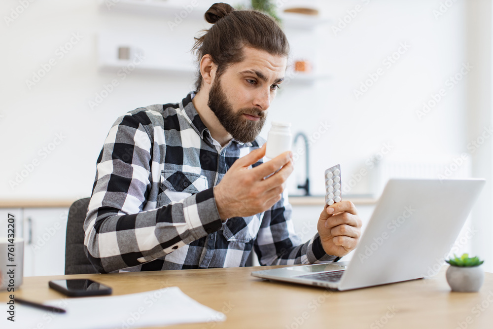 Fototapeta premium Medicine containers held by Caucasian young man looking through information on laptop screen in kitchen. Remote employee suffering from stuffy nose during allergy season at work from home.