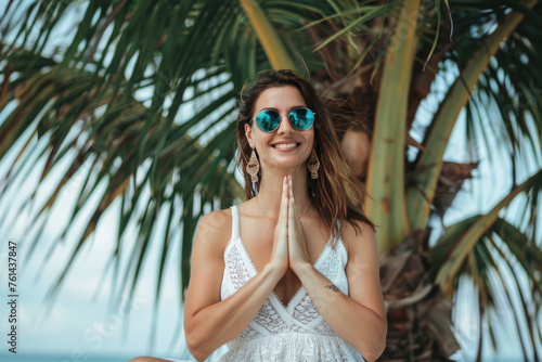 Beautiful young woman doing yoga on palm tree at tropical beach, happy and smiling. Beauty fashion model in white summer with blue sunglasses sitting crosslegged isolated over bright sunny sky backgro