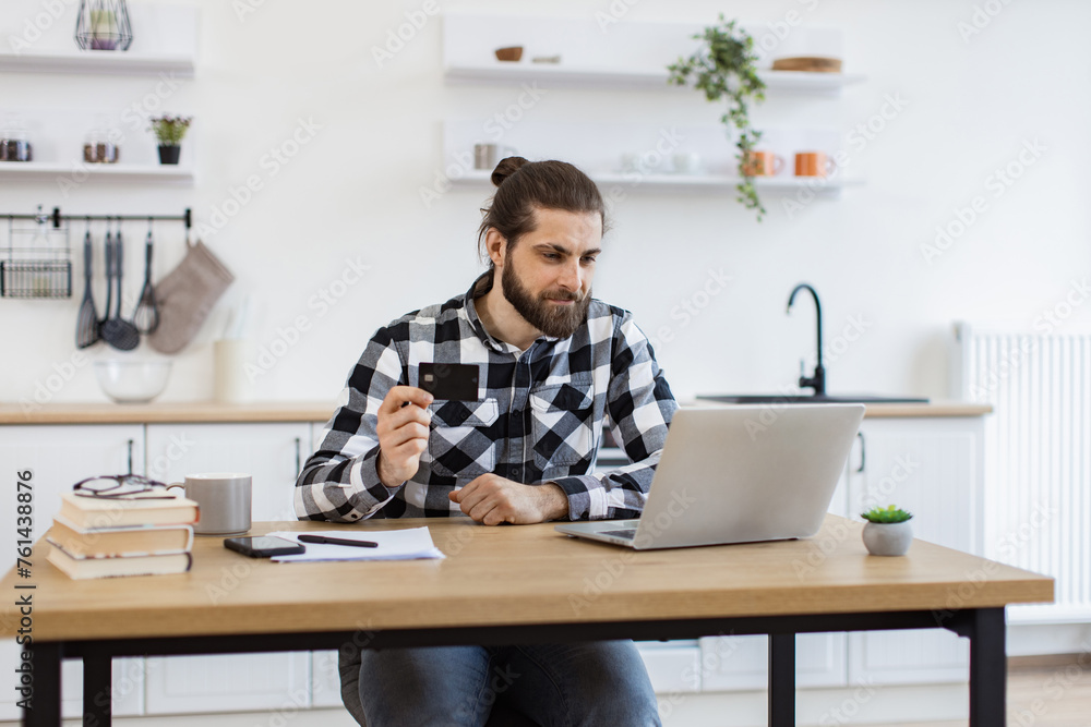 Joyful Caucasian man in casual clothes using credit card while running portable computer in workplace. Smiling bearded person in checkered shirt enjoying virtual shopping using online wallet from home Joyful Caucasian man in casual clothes using credit card while running portable computer in workplace. Smiling bearded person in checkered shirt enjoying virtual shopping using online wallet from home