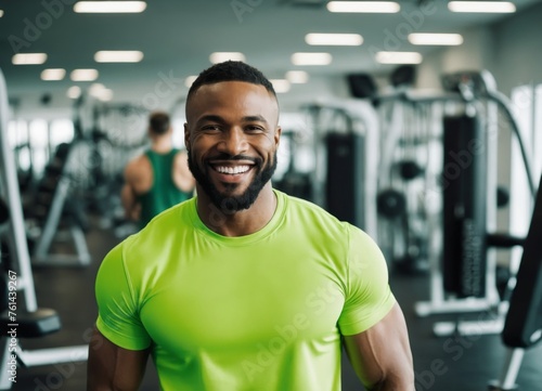 Muscular African American man in green sportswear, fitness trainer smiling and looking at the camera on the background of the gym