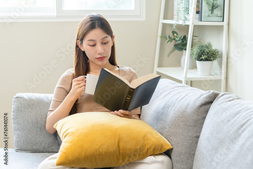 Having relaxing weekend, comfort asian young woman with cup of beverage, drinking coffee or tea of mug. Girl sitting on sofa open reading book enjoy of rest after get up in morning lifestyle at home.