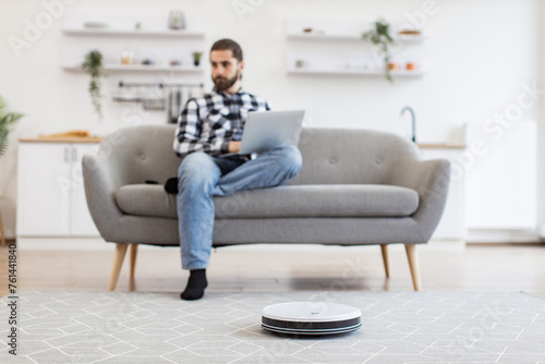 Cheerful owner programming home gadget via remote control while focusing on work. Focus on smart robotic vacuum operated by happy Caucasian man while performing automatic cleaning on carpet indoors.
