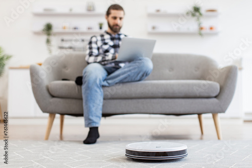 Cheerful owner programming home gadget via remote control while focusing on work. Focus on smart robotic vacuum operated by happy Caucasian man while performing automatic cleaning on carpet indoors. © sofiko14