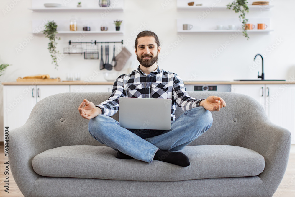 Peaceful Caucasian businessman in casual attire sitting on couch with wireless laptop on knees and meditating. Bearded young guy with fingers in mudra gesture relieve stress and regaining energy. - obrazy, fototapety, plakaty 