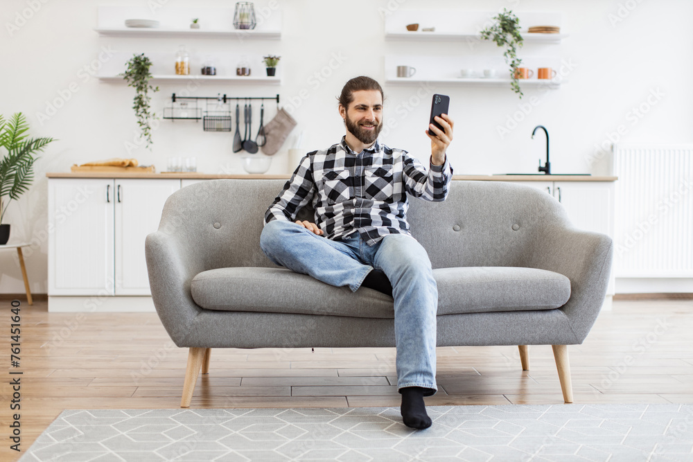 Attractive young man holding phone while talking via webcam on kitchen. Happy Caucasian person in checkered shirt conducting online conference via modern device sitting on couch in home interior.