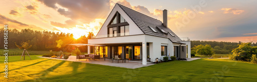 Photo of modern house with gable roof and terrace on the first floor, white walls with black window frames in an environment surrounded by green grass at sunset © Kien