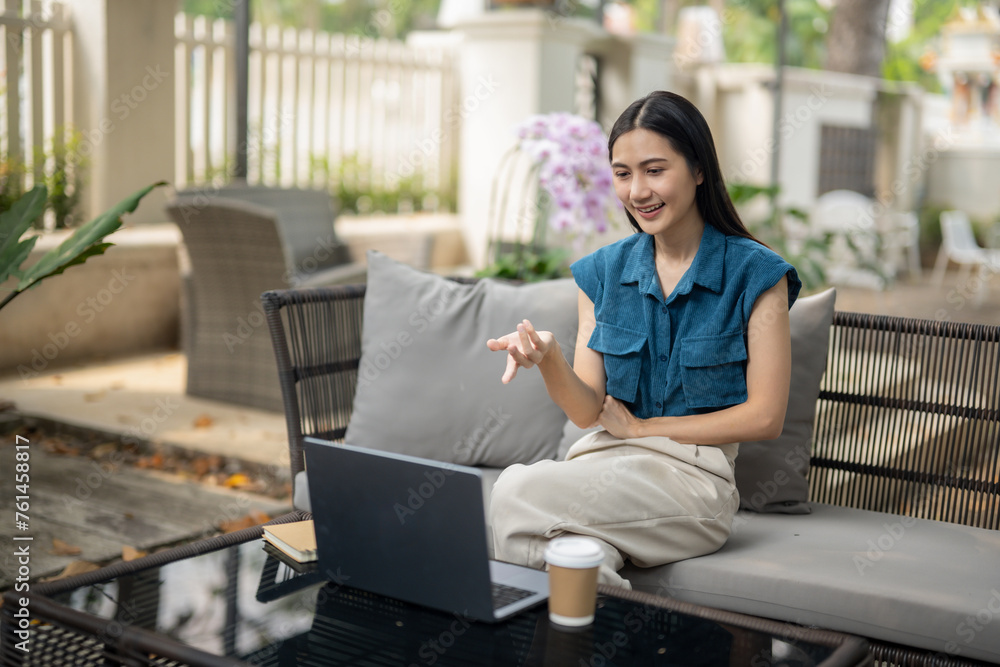 Young Asian woman having a video call on her laptop. While sitting on a sofa in the garden of a cafe with a cup of coffee. Young Asian woman having a video call on her laptop. While sitting on a sofa in the garden of a cafe with a cup of coffee.