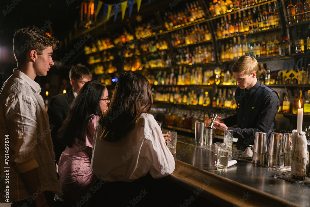 Young customers wait for cocktails sitting at bar counter. Experienced bartender concentrates on making alcoholic drinks for visitors