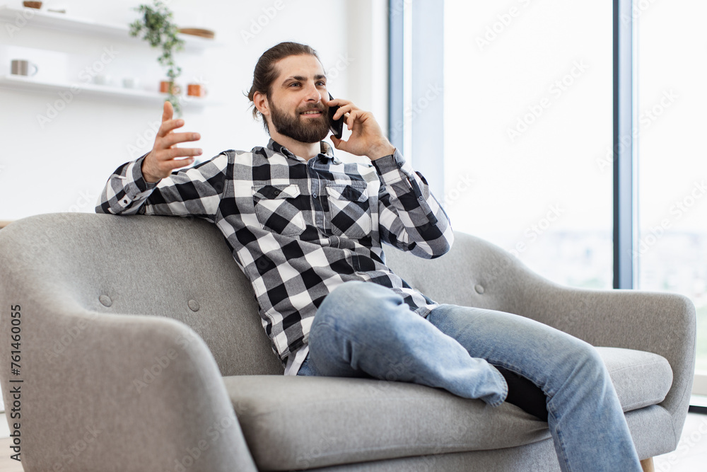 Side view of smiling business person sitting on couch while chatting on cell phone in bright room on sunny day. Professional Caucasian manager discussing report at light open plan kitchen at home. Side view of smiling business person sitting on couch while chatting on cell phone in bright room on sunny day. Professional Caucasian manager discussing report at light open plan kitchen at home.