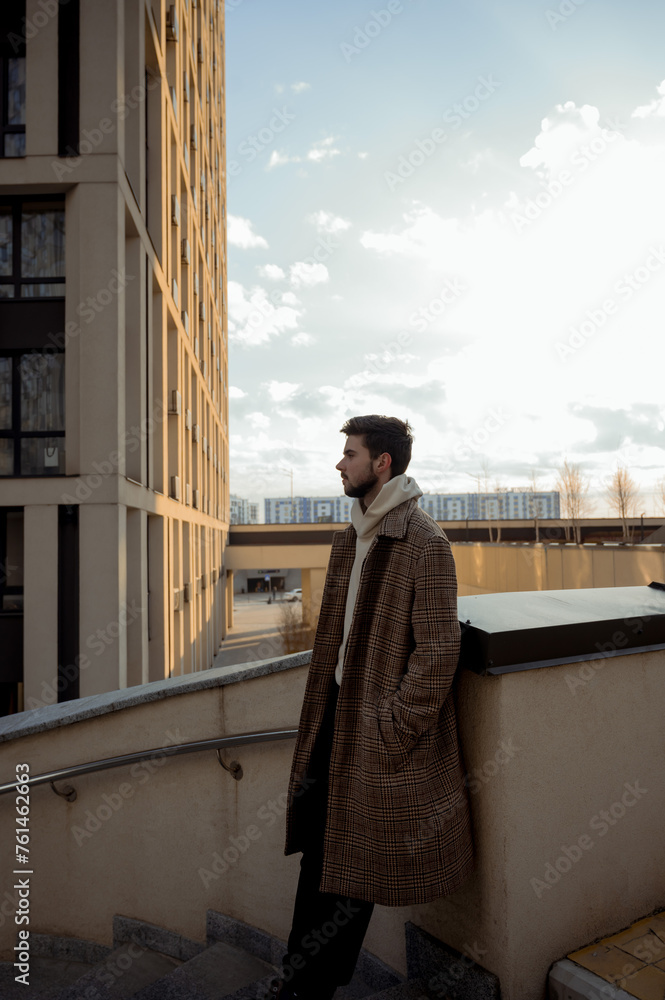 A stylish man in a coat stands on a city street with a modern building in the background. young guy standing in a city block.