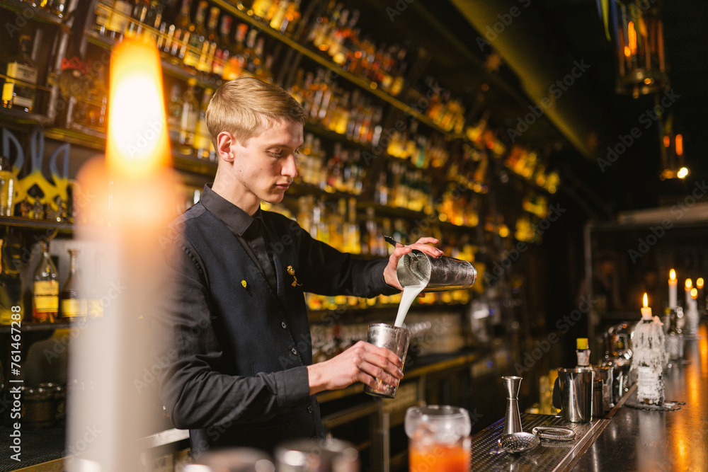 Bartender pours alcoholic milkshake from one part of shaker to another to froth drink. Barman stands by bar counter background antique-style shelves