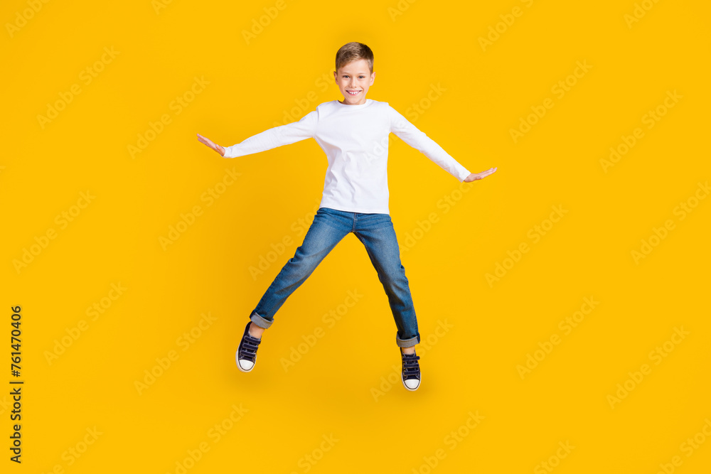 Full length photo of carefree positive kid dressed white shirt arms sides jumping high empty space isolated yellow color background Full length photo of carefree positive kid dressed white shirt arms sides jumping high empty space isolated yellow color background