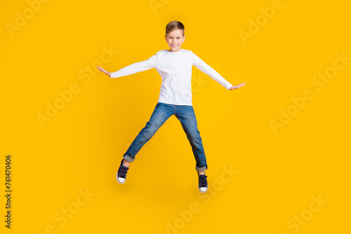 Full length photo of carefree positive kid dressed white shirt arms sides jumping high empty space isolated yellow color background
