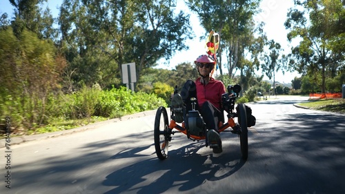 Elderly senior woman riding recumbent tricycle e-bike towards the camera on a path on a sunny day in a park.