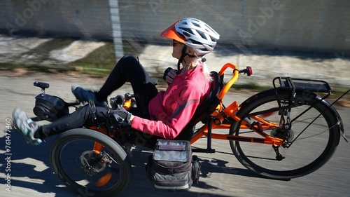 Rear side view of elderly senior woman riding recumbent tricycle e-bike on a path on a sunny day in a park.