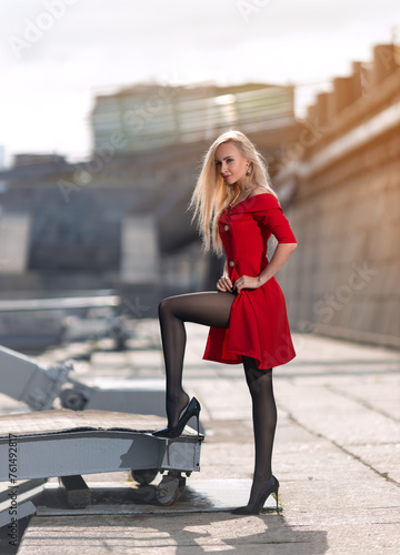 Beautiful blonde woman in a red dress with perfect legs in pantyhose posing outdoors on the autumn street in the lights of the setting sun.