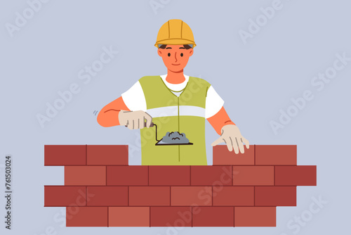 Man bricklayer builder builds brick wall using trowel with concrete mixture to secure blocks. Guy bricklayer in yellow vest and hardhat works in architectural company performing contract work