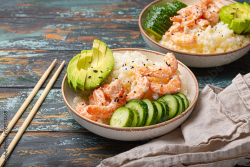 White ceramic bowl with rice, shrimps, avocado, vegetables and sesame seeds and chopsticks on colourful rustic wooden background front view. Healthy asian style poke bowl.
