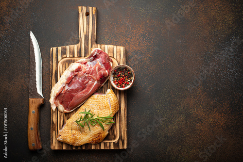 Two raw uncooked duck breast fillets with skin, seasoned with salt, pepper, rosemary top view on wooden cutting board with knife, dark brown concrete rustic background, space for text.