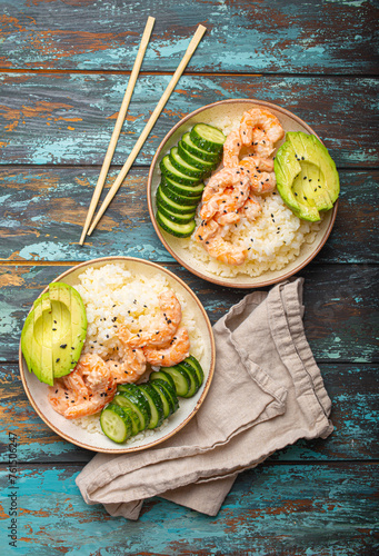 Two white ceramic bowls with rice, shrimps, avocado, vegetables and sesame seeds and chopsticks on colourful rustic wooden background top view. Healthy asian style poke bowl.