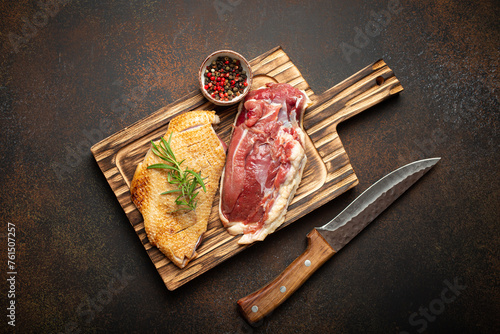 Two raw uncooked duck breast fillets with skin, seasoned with salt, pepper, rosemary top view on wooden cutting board with knife, dark brown concrete rustic background.