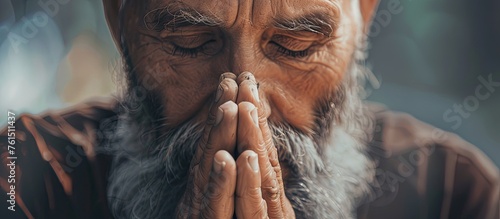 A man with facial hair and a beard is making a praying gesture with his hands folded in front of his face. The wrinkles on his forehead show his concentration photo