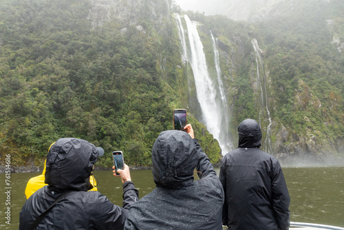 Milford Sound, Fiordland, New Zealand _ Nov. 14, 2023: Tourists snap pictures of a magnificent waterfall froim a ferry on Milford Sound, one of the wettest places on the planet. photo