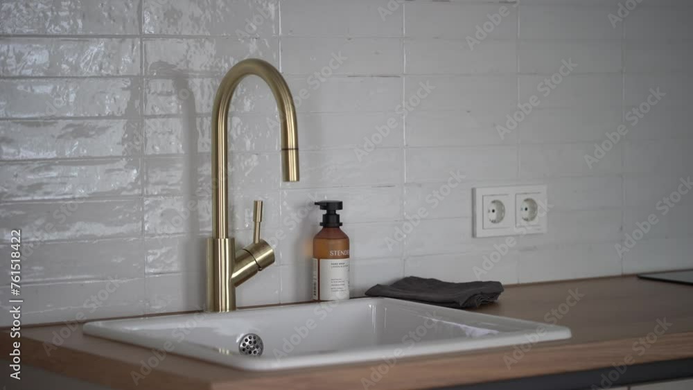 Cesis, Latvia - February 14, 2024 - a kitchen sink with a modern gold faucet, beside a soap dispenser and a cloth, set against a tiled backsplash with electrical outlets. Cesis, Latvia - February 14, 2024 - a kitchen sink with a modern gold faucet, beside a soap dispenser and a cloth, set against a tiled backsplash with electrical outlets.