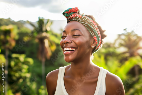 A beautiful young black woman with a colorful headband smiles and laughs with her eyes closed. She has a white T-shirt. In the background is a tropical garden with palm trees