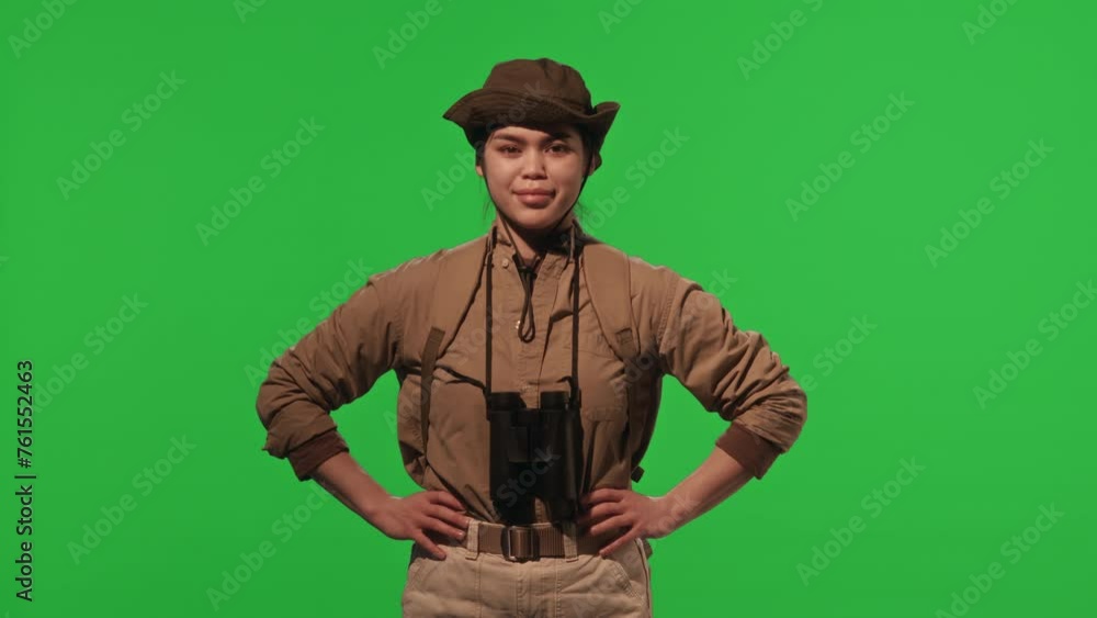 Portrait of young female tourist with backpack and binoculars holding hands on hips and posing for camera with confidence against chroma key green background Portrait of young female tourist with backpack and binoculars holding hands on hips and posing for camera with confidence against chroma key green background