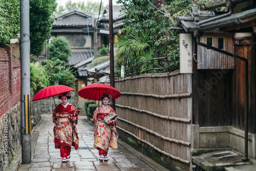 Geishas navigate the wet cobblestones of Kyoto in their radiant attire, the red umbrellas a poetic touch to the grey skies. They move with a grace that honors their cultural legacy photo