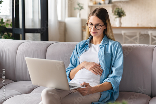 Young caucasian pregnant woman sitting on the sofa and using laptop. Baby expectation. Searching browsing information and maternity leave. Social distance concept