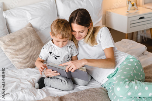 Loving caring young mother reading a book with her little toddler son lying on a bed together. Weekend pastime with family. Stay at home, children care concept