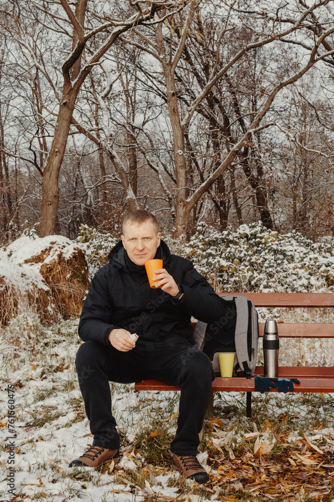 Fototapeta premium Winter Serenity: 40-Year-Old Man Enjoying Tea on Snow-Covered Bench in Rural Park. Immerse yourself in the tranquil beauty of winter as a 40-year-old man finds solace on a snowy bench in a rural park.