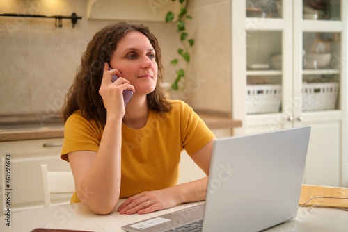 A woman with a laptop is talking on a mobile phone while sitting at a table in a home kitchen. An adult female businesswoman works from home, a remote office