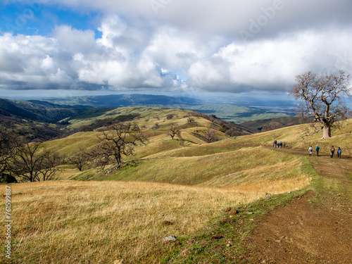 A group of hikers descending a trail along a mountain ridge into a distant valley with large oak trees dot grass covered hills, Ohlone Wilderness, California
