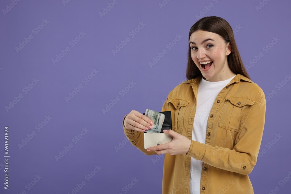 Excited woman putting dollar banknotes into wallet on purple background, space for text Excited woman putting dollar banknotes into wallet on purple background, space for text